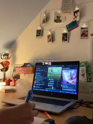 A well-lit room with a desk, a laptop, and some decorations. The laptop is open and displaying a screen, likely for studying. There are some photos and lights on the wall.