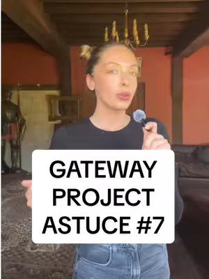 A woman in a black top and jeans stands in front of a background featuring a red wall, a doorway, and a chandelier. She's holding a microphone and a white sign that has the words "GATEWAY PROJECT ASTUCE #7" in bold, black text. The overall shot appears to be from a casual video.