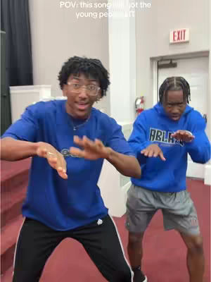 Two young men are captured in a medium shot, posed with their hands down, seemingly dancing or mimicking a song. They wear casual outfits, with matching blue tops and contrasting bottoms, and are positioned in what appears to be a church interior, framed by red carpet along the bottom and a white wall in the background.