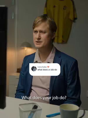 A man wearing a blue blazer and a pink shirt is sitting at a desk and looking at something off screen. A speech bubble lies next to his upper body and it seems to be a comment someone left. In the background there is a soccer/football jersey hanging on the wall.