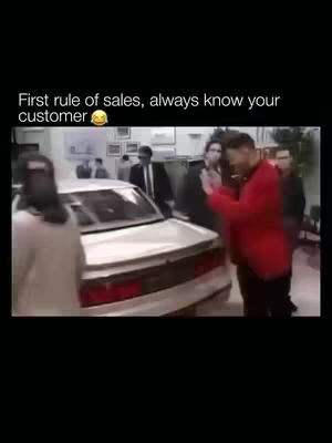 The image is a screenshot from a movie or show. It depicts a man in a red blazer approaching a car, with several other people, some in suits, standing nearby against a backdrop of what may be an office or dealership setting.