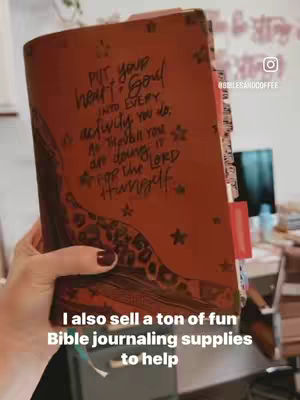 A hand holding a Bible that is open toward the viewer. The cover is brown with various words written on it in cursive and there are several star embellishments. The hand has dark red nail polish. Below the Bible is text in white font. The video is taken in front of a desk or a space that appears to be a workspace.