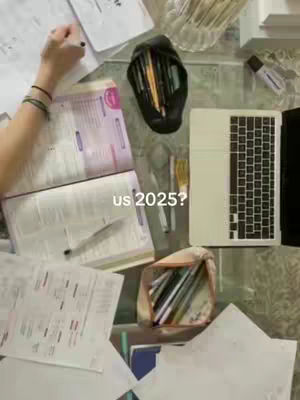 An overhead shot of a cluttered glass table filled with study materials. In the center is a book, papers, and a laptop. A hand, presumably the student's, is visible on the left side with a pen in hand. Scattered pens, pencils, and other stationery items complete the scene, implying a study environment.