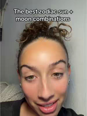 A close-up shot of a woman's face with a background of white and a hint of cream. The text overlay says: "The best zodiac sun + moon combinations." The woman has light skin, curly brown hair pulled back, and is smiling slightly with reddish lipstick. She has two small earrings and is in a black top.