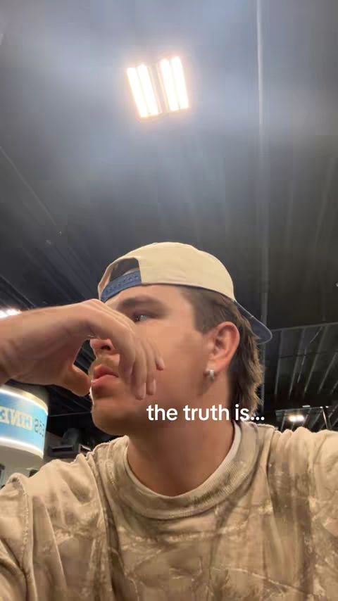 A young man, viewed from the chest up, gazes downward with a questioning expression. He has short, brown hair styled in a slight mullet and a light beige baseball cap with a dark navy brim. He wears a tie-dyed earth-tone shirt, and his hand is raised near his face. The background, out of focus, reveals a glimpse of a commercial interior lit by large fluorescent lights.