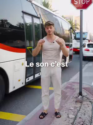 A man stands in front of a bus, gesturing with his hands. The text overlay says "Le son c'est." The scene appears to be outdoors on a sunny day.
