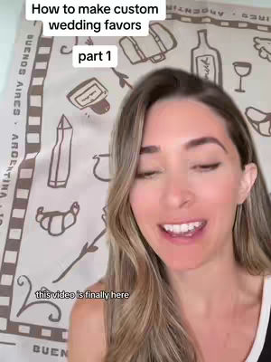A woman smiles directly at the camera. Behind her, a light-colored fabric with a pattern of sketched wedding-related images like luggage, bottles, and a wine glass serves as a backdrop. The overlaid text reads, "How to make custom wedding favors" across the top, with "part 1" below it, and "this video is finally here" at the bottom. The woman has long hair and wears a white top.