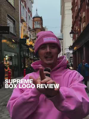 A man stands in a city street with a pink outfit. The hook overlaid onto the image states "SUPREME BOX LOGO REVIEW". He wears a pink hoodie and a beanie, also labeled with a supreme logo. He is speaking into a microphone.