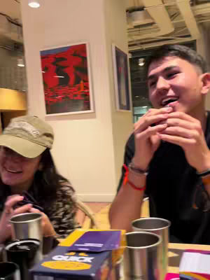 A cover image captures a scene of friends around a table, engrossed in a board game. Two young adults are the main features. The male on the right is mid-laugh with his hands up. The female, on the left, is smiling, wearing a cap. A board game box and metal cups are visible, and a painting hangs on the wall.