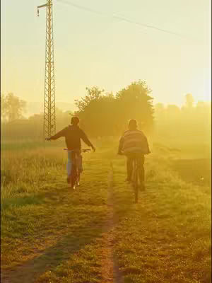 Two people riding bicycles on a dirt path through a grassy field during a golden sunset. The sky is bright yellow with the sun behind the riders. A tall, metal pylon is on the left side of the image. The riders are silhouetted, making it impossible to identify their features.