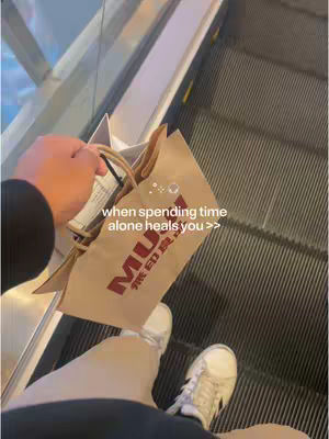 A first-person view from an escalator, showing the creator's perspective. A person is holding a MUJI shopping bag with a partially visible hand; below this are the person's legs and feet with white sneakers. Escalator steps flow down in the background. The image has a low-angle perspective, focusing on retail and personal time.