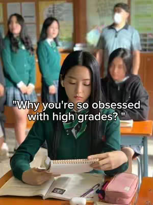 A young woman, centered in the image, sits at a desk and looks down at an open notebook. Additional people are blurred in the background, suggesting a school setting. The primary colors are various shades of green and neutral office colors.