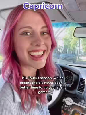 A woman with pink hair smiles widely while driving a car. Text overlays appear at the top saying 'Capricorn' and at the bottom, text that reads, 'It's Taurus season, which means there's never been a better time to up your scent game.' The steering wheel and car interior are visible, and a Febreze car air freshener is in view.