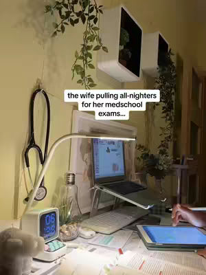 A cozy desk setup with a laptop, tablet, and books. A stethoscope hangs on the wall. Text overlay: 'the wife pulling all-nighters for her medschool exams...'