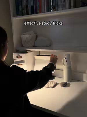 A dimly lit image shows a person from the back at a desk, possibly studying. At the top, a shelf is visible with books and decorative items. The person's hand is interacting with a tablet on the desk. The desk surface is white, with a keyboard, mouse, water bottle, and pen visible. The main text overlay reads: "effective study tricks".