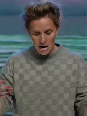 A woman with short, tousled hair and a checkered grey top is the central figure, looking down with her mouth open. The background is blurred, with hints of green and blue, suggesting an indoor setting. The lighting appears to be soft and even.