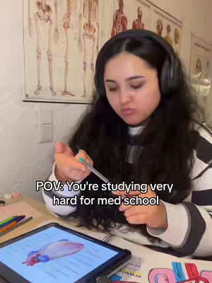 A young woman wearing headphones is seated at a desk. She is holding a pen and appears to be studying, with a digital tablet displaying a diagram of a heart in front of her. Anatomical charts are visible in the background.