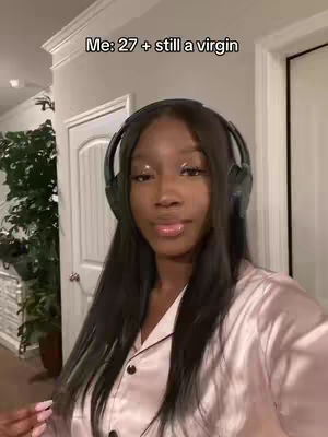 A woman with long black hair and headphones is centered in the frame against a neutral background. Text overlays stating her age and virginity. She's wearing a light pink satin shirt and appears to be indoors.