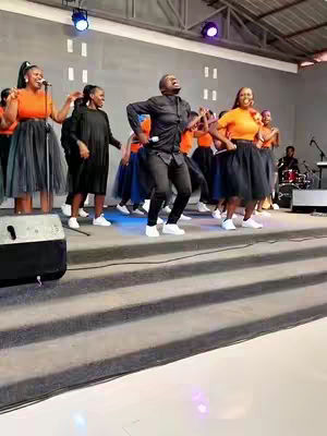 A group of people are on a stage, with a man in the center who appears to be leading a performance. Two other women are holding microphones.