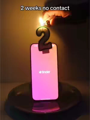 The image shows a smartphone with the Tinder app open, placed on a dark surface. A candle shaped like the number "2" is lit above the phone. The background is a gradient. The lighting suggests a warm, intimate setting.