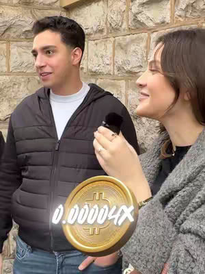 A man and a woman are standing in front of a stone wall. The man on the left is wearing a black jacket, a white shirt, and blue jeans. The woman on the right is wearing a gray sweater and holding a microphone. A gold coin with a Bitcoin symbol and the value 0.00004x is digitally overlaid in the image.