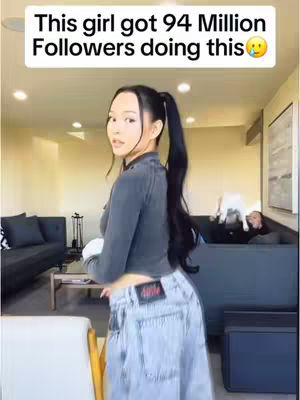 A young woman stands in a modern living room, looking back over her shoulder. She wears a dark top, jeans, and has long, dark hair in a high ponytail. Text in a rectangular box at the top says: 'This girl got 94 Million Followers doing this' with a surprised face emoji. The background includes a couch, white walls, and windows, suggesting a casual home environment.