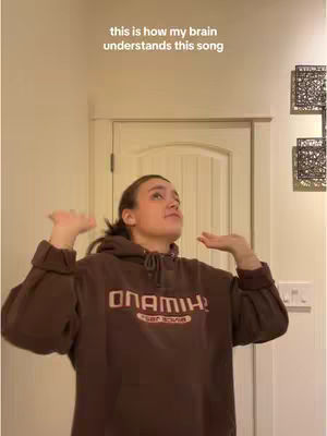 A young woman stands in a room, arms raised and palms upward, seemingly reacting to a song. She wears a brown hoodie with text on it. The background includes a closed white door and decorative wall art.