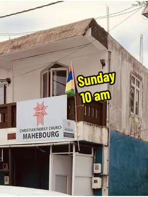 An exterior shot of a two-story building, likely a church, with a sign that reads "Christian Family Church MAHEBOURG." The building is white with a brown roof and features a balcony with a small flag. Overlaid on the image is text in a yellow bubble that says. The overall composition suggests a promotional image for a religious service.