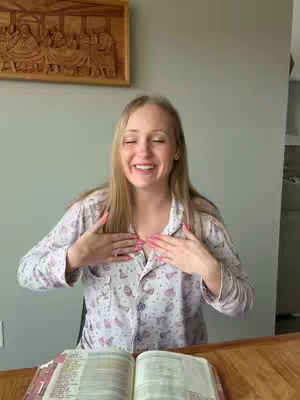 A woman with long blonde hair and a pink floral print pajama shirt is smiling with her eyes closed, hands on her chest, over a Bible partially open on a wooden table, with a wooden carving in the background.