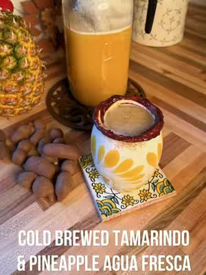 The cover image displays a close-up shot of a mixed drink sitting on a wooden surface. The drink is in a tall glass with a light orange hue. Alongside the glass, there's a small, decorated cup with a dark brown liquid inside, placed on a patterned coaster. Whole tamarind pods are scattered to the left of the drink. There is a pineapple partly visible on the top left.