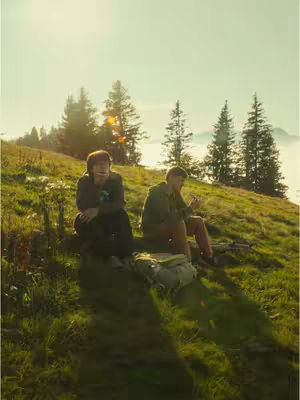 Two young men are sitting on a grassy hillside, surrounded by green vegetation and tall pine trees. The sky is a soft, pale yellow, suggesting either early or late daylight. The men are dressed in casual attire, and the scene conveys a sense of relaxation and natural beauty.
