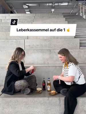 Two young women are sitting on concrete steps, smiling and appearing to prepare food and drink. One is opening a red bottle, the other is holding a sandwich. There are bottles and sandwich. The background is a plain concrete wall.