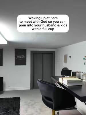 A room interior with a desk, chair, and inspirational text overlay. The setting is a home office or study, with neutral walls, a dark desk, and a black office chair. A text overlay at the top reads, 'Waking up at 5am to meet with God so you can pour into your husband & kids with a full cup'. The overall composition is simple and focuses on the text and the implied activity of the space for reflection and work.
