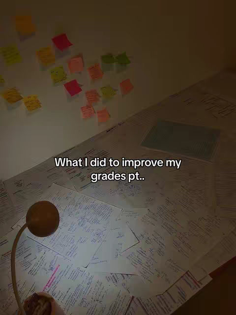 The image shows a messy desk with papers strewn across it. A table lamp with a round, brown head sits in the foreground, casting a warm light. Colored sticky notes of different sizes and colors are stuck on the wall in the background. The text overlay reads, "What I did to improve my grades pt.."