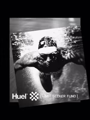 A black and white image of a person swimming underwater. The swimmer is seen from the chest up, with arms extended and a determined expression. Water and bubbles surround the swimmer, and the image is framed with elements giving it a layered look. The Huel logo and text are at the bottom.
