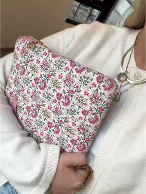 A person is holding a patterned bag with a floral design, wearing a white sweater and necklaces, appearing to be the central focus of the image.