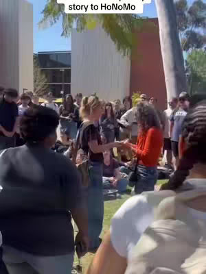 An outdoor scene shows a crowd gathered around two women. One, in a red top, appears to be talking to the other who is holding her hands. The background includes people of various ethnicities and ages, suggesting a public space or event at a university.