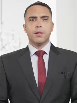 A medium shot featuring a man in a dark suit and a red tie. He is positioned centrally and looking directly at the camera. The background is a soft, out-of-focus white, suggesting a studio setting.