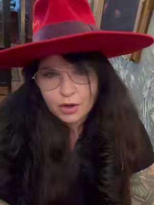 A woman with long black hair, glasses, and a red hat is centered in the frame, looking directly at the camera and speaking. The background is a blend of indistinct patterns and colors, suggesting an interior setting.