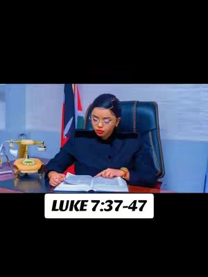 A woman with glasses is seated at a desk, reading a book. She's wearing a dark blazer and a gold watch. A Kenyan flag is visible in the background, along with a desk lamp. Text in white on a black rectangle at the bottom reads: 'LUKE 7:37-47'. The background is dark.