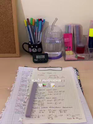 A still life setup of a desk with study materials. A notepad with handwritten notes and a pen holder filled with colorful pens are in focus. There's also a water bottle, timer, and other stationery items on the table.