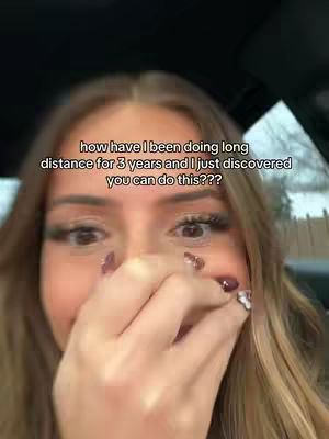 The cover image is a selfie featuring a young woman. The woman is fair-skinned, with long, blonde hair, and is holding her right hand up to her mouth, partially covering it. Her prominent, well-defined eyebrows and long eyelashes are visible, and she's wearing makeup. The background is blurred, likely inside a car. The text overlay reads: "how have I been doing long distance for 3 years and I just discovered you can do this???"
