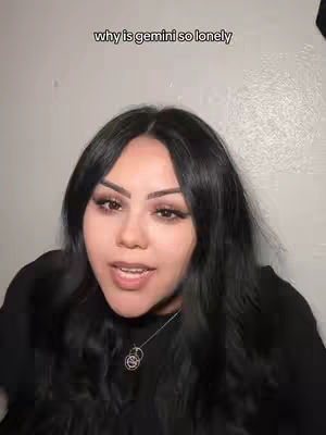 A woman with long, dark hair is centered in the frame, facing the camera. She has full makeup on, including eyeshadow and lipstick. In the background, a plain wall serves as a backdrop. The image is well-lit, creating a clear focus on the woman as she speaks to the viewer.