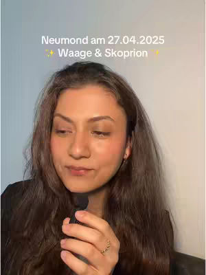 A woman with long, dark hair is centered in the frame, holding a microphone. Text overlays the image indicating a new moon on a specific date, with astrological signs.