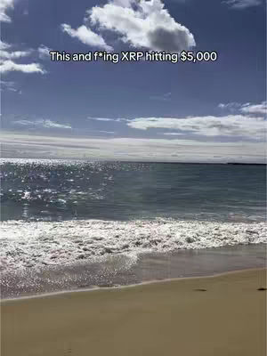 A serene beach scene with a bright blue sky dotted with fluffy white clouds. The ocean, a mix of deep blue and shimmering light, meets a sandy beach in the foreground. The composition is balanced, with the sky taking up a significant portion of the frame and the beach providing a grounding element.