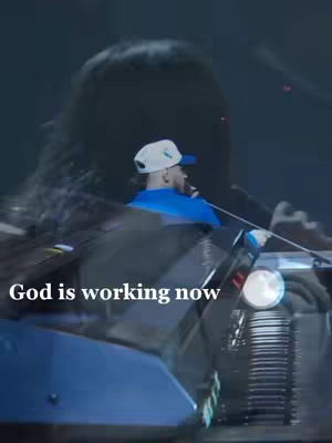 The cover image shows a man in a blue shirt and a white cap, illuminated by stage lights. A larger, blurry silhouette of another person hovers in the background. The text 'God is working now' is overlaid in white at the bottom half of the image.