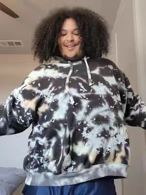 A person with curly dark hair is wearing a tie-dye hoodie, standing with arms out against an indoor background. The person is smiling.