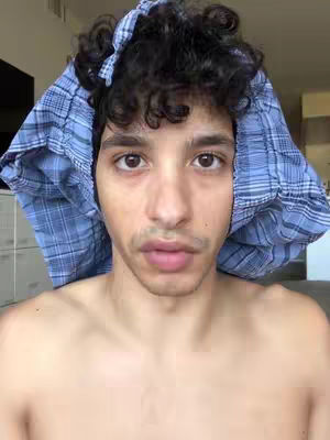 A man is centered with a blue patterned cloth over his head, looking directly at the camera. The image is a selfie, with a plain background suggesting an indoor setting. The man's facial expression is neutral.