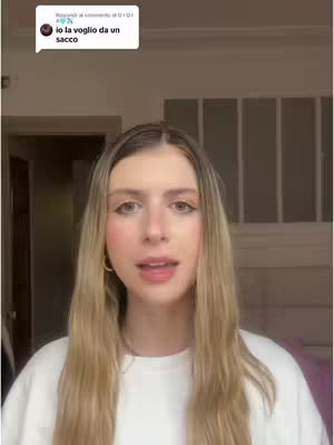 A young woman with long blonde hair looks directly at the camera, speaking. She wears a white sweater. Behind her, a simple, well-lit room with a window and wooden elements provide a neutral backdrop.