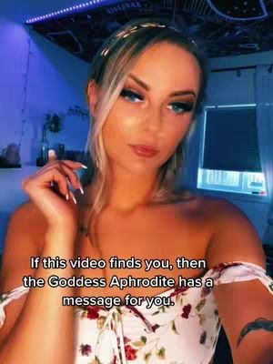 A woman with long blonde hair and a gold headband is centered in the frame. She has makeup on, including dark eyeliner and pink lip color and wears an off-the-shoulder floral top. Text overlayed on bottom of the image. The background has cool blue lights and glimpses of a window.